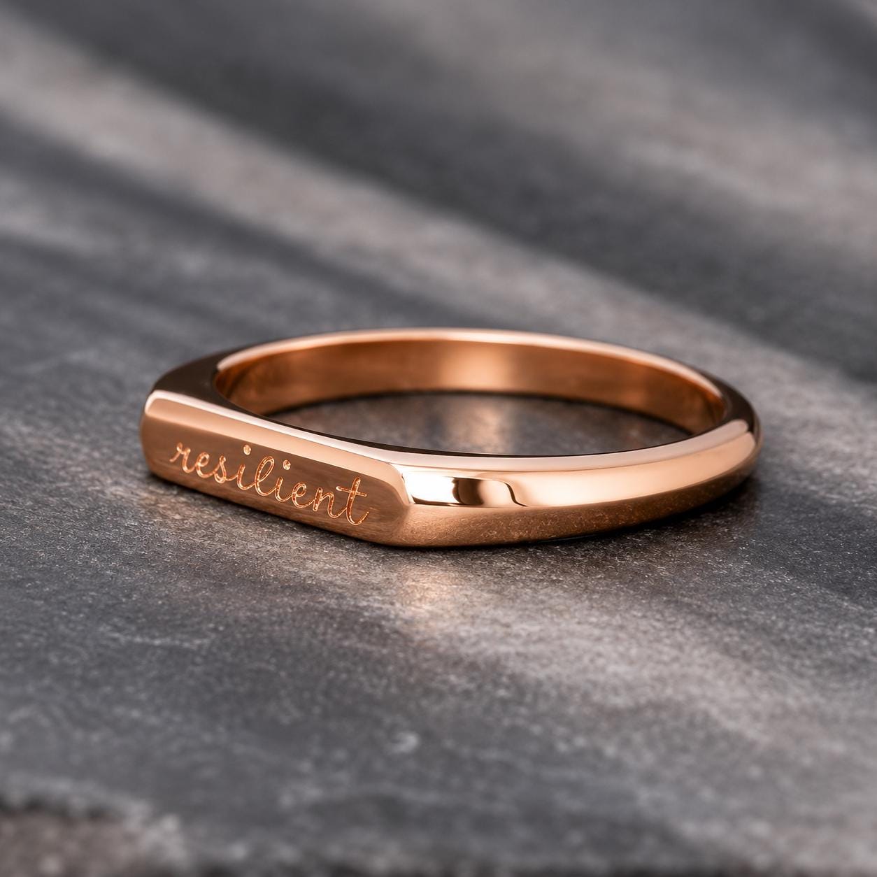 A rose gold ring with the word "resilient" engraved on it, resting on a gray surface.
