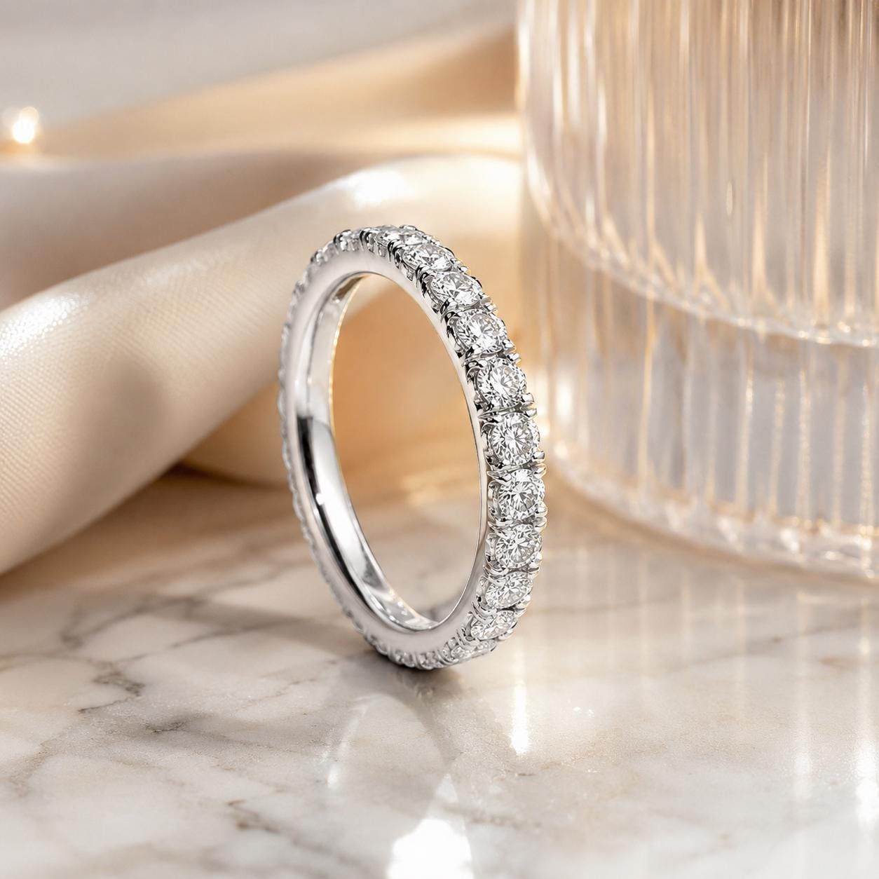 A diamond-encrusted ring with a row of diamonds set in a band is displayed on a marble surface, with a glass object in the background.