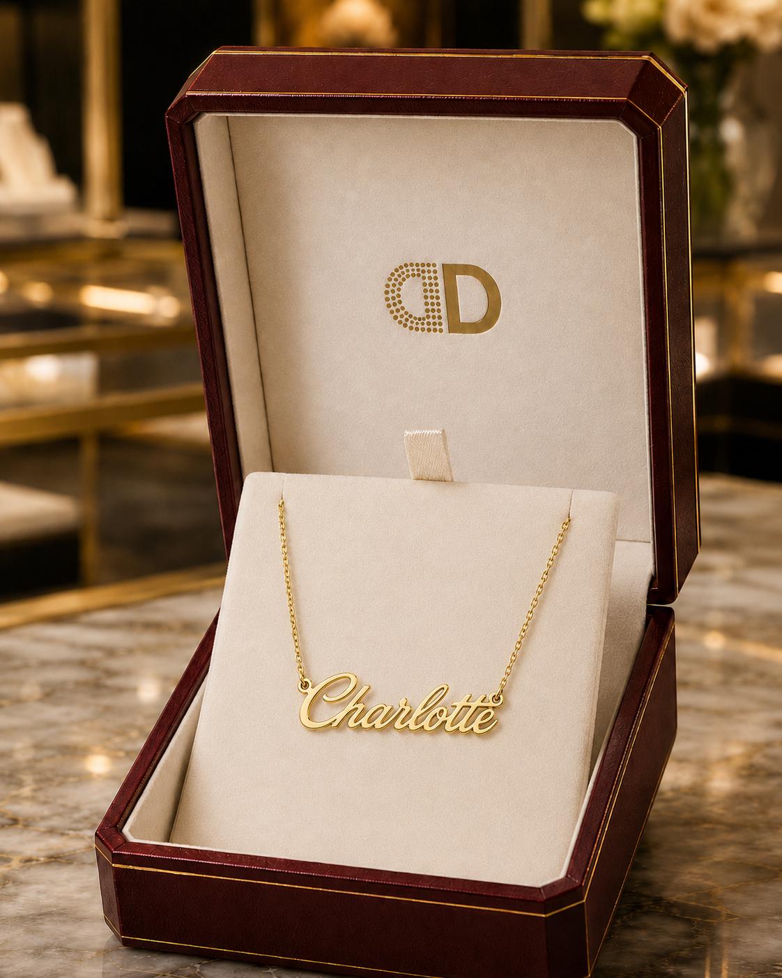 A gold necklace with the name "Charlotte" engraved on a white stand, placed in a burgundy jewelry box with a gold logo.