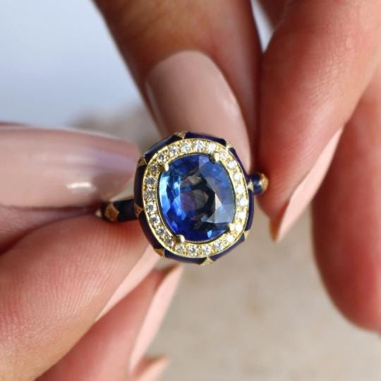 A close-up image of a person's hand holding a ring with a large blue gemstone surrounded by smaller diamonds.