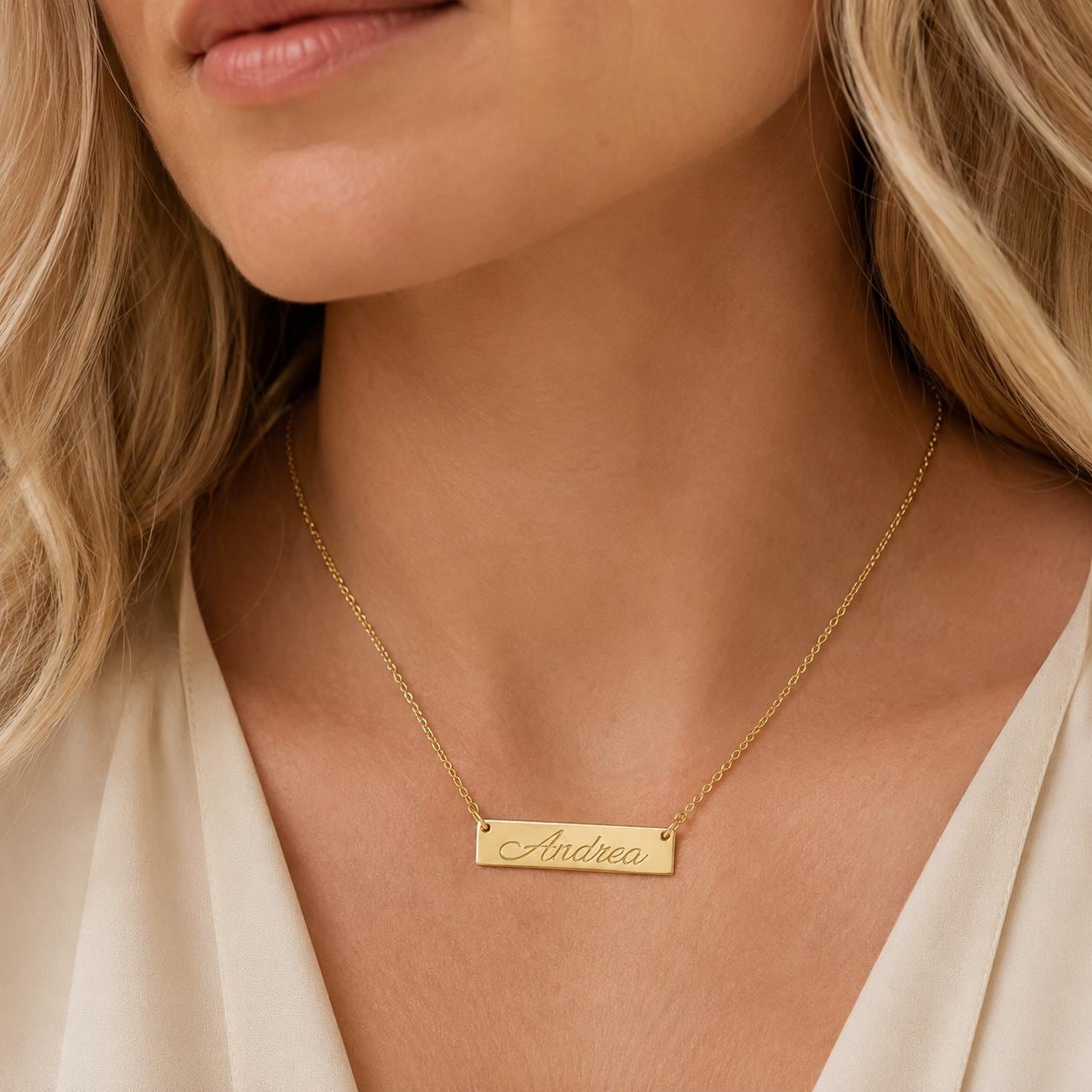 A close-up of a woman's neck wearing a gold necklace with a rectangular pendant engraved with the name "Andrea".