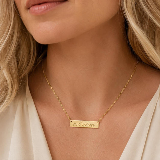 A close-up of a woman's neck wearing a gold necklace with a rectangular pendant engraved with the name "Andrea".