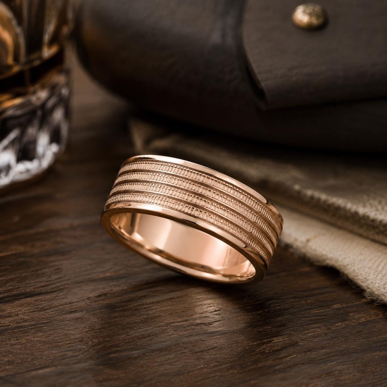 A gold ring with a textured band is displayed on a wooden surface, with a glass and a leather item in the background.