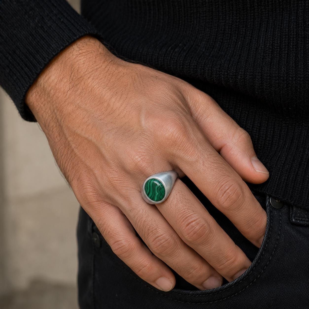 A close-up of a person's hand wearing a ring with a green stone, resting on their hand near their pocket.