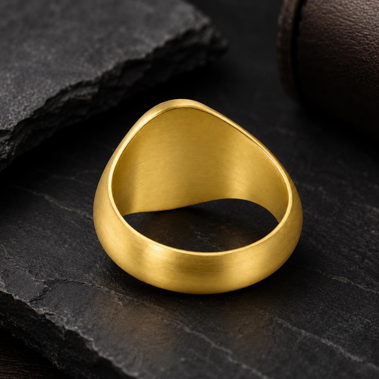 A gold ring with a unique design sits on a dark surface, surrounded by a few other dark objects.