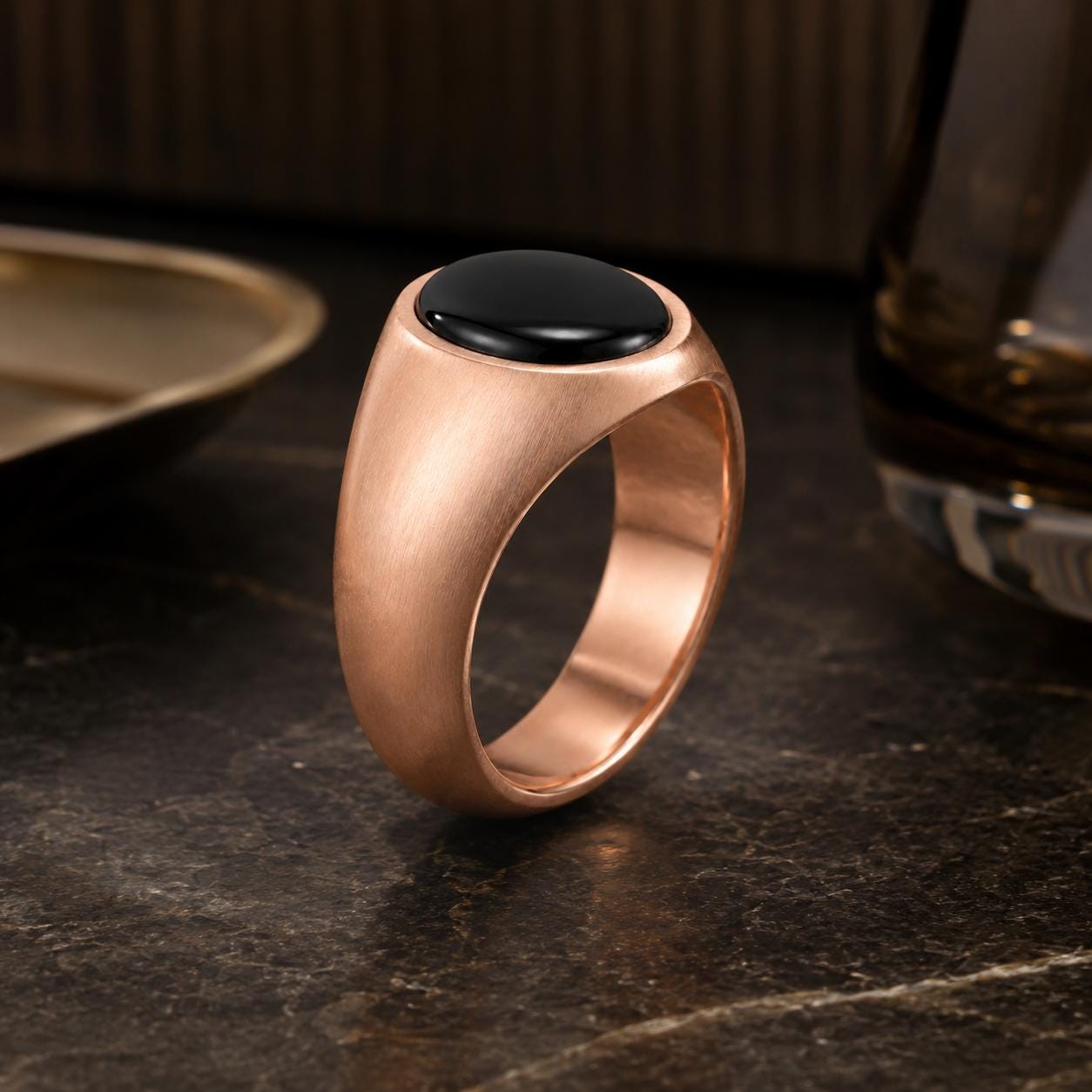A gold-colored ring with a black gemstone sits on a dark surface, with a glass and a plate visible in the background.