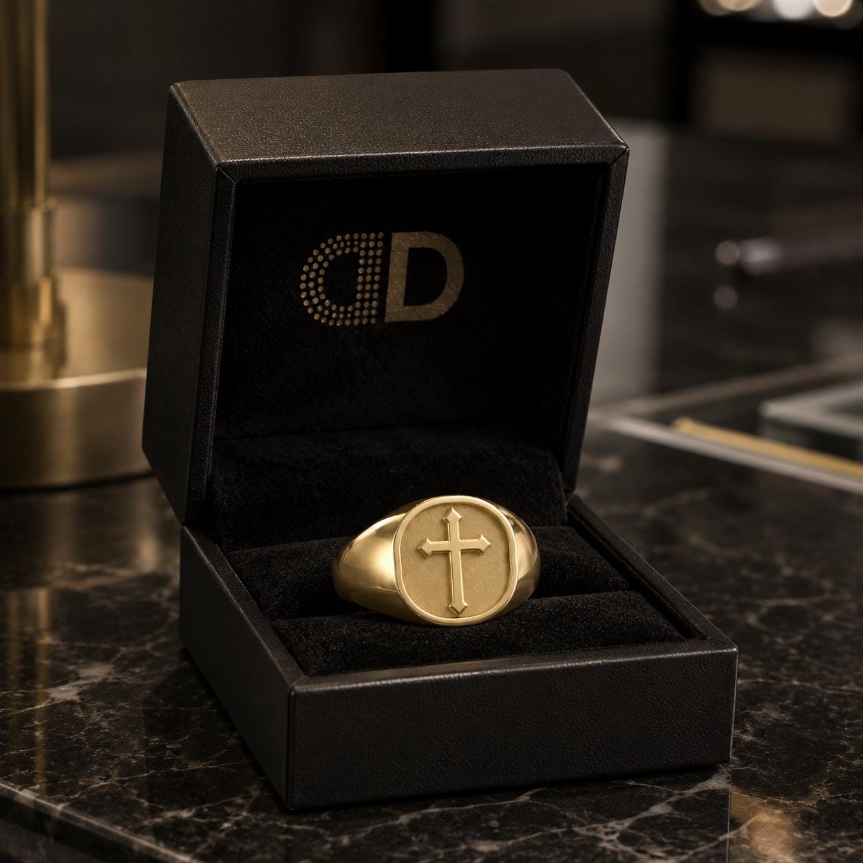 A gold signet ring is displayed in a black jewelry box with a gold logo on the lid.