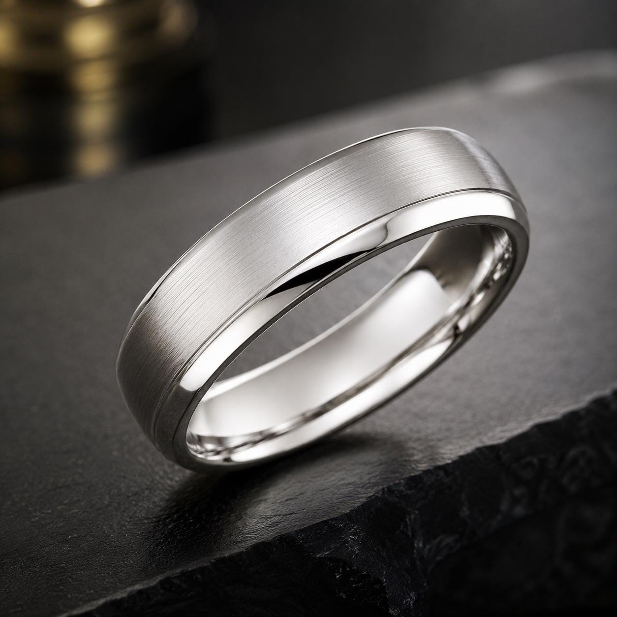 A silver ring with a smooth, polished surface sits on a dark surface, possibly a stone or a black table.