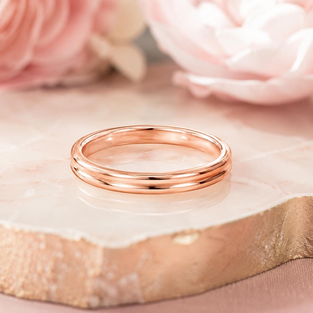 A gold-colored ring is displayed on a marble surface, with a pink flower in the background.