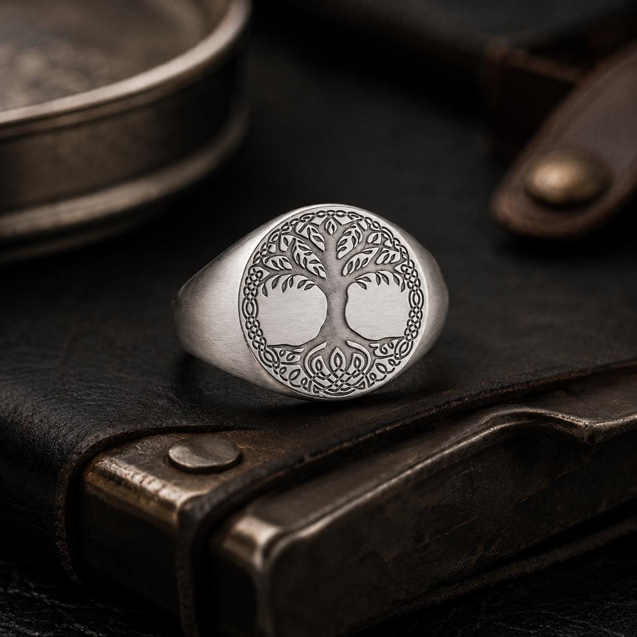 A silver ring with an intricate tree design sits on a leather surface, surrounded by other objects.
