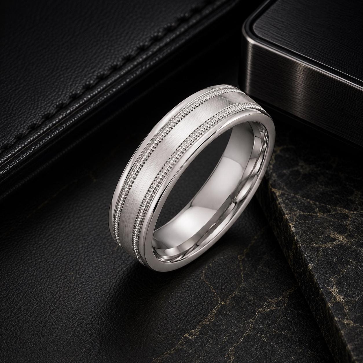 A silver ring with a textured band is displayed on a black surface, with a black leather-like object partially visible in the background.