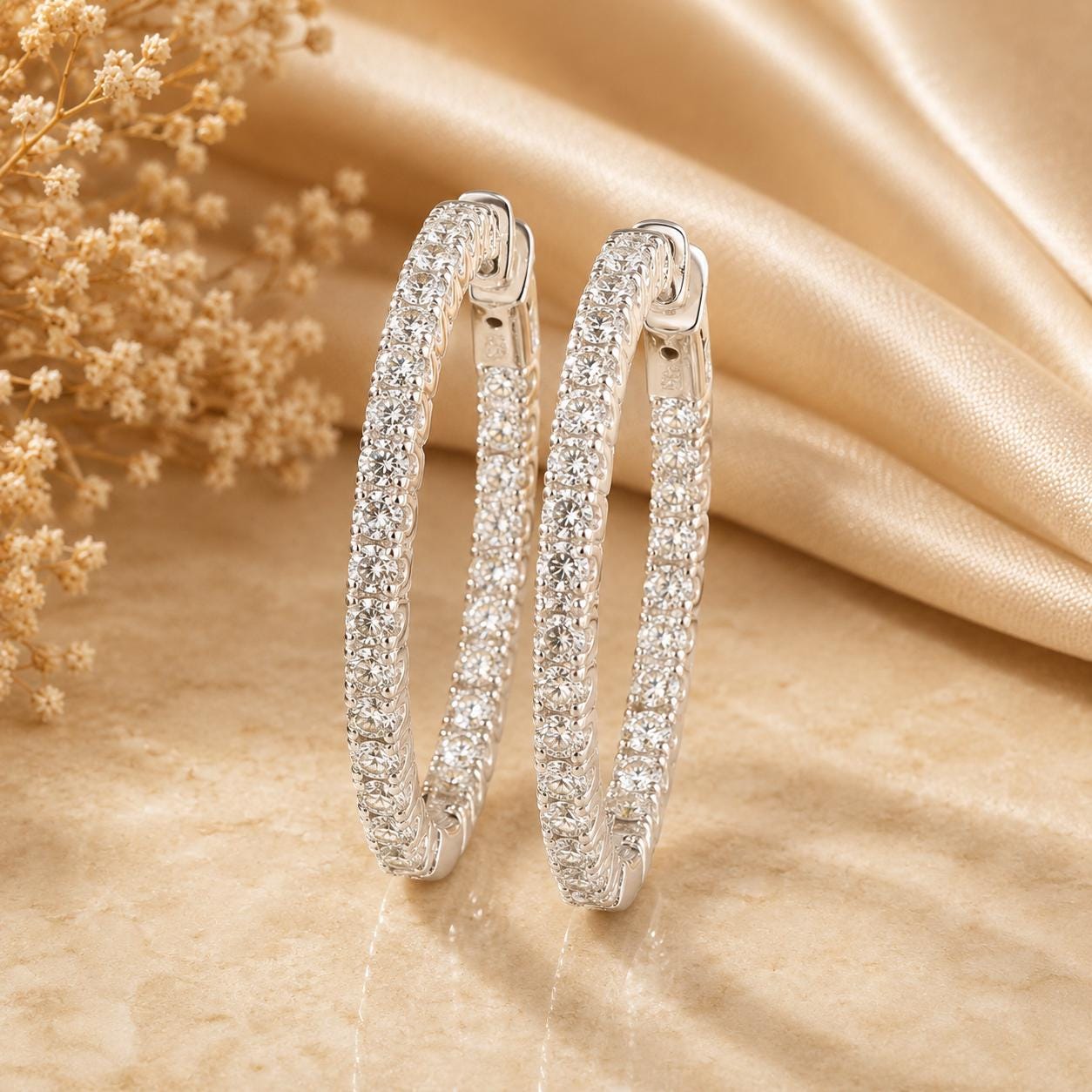 A pair of elegant diamond-encrusted earrings is displayed on a beige fabric background, with delicate white flowers visible in the background.