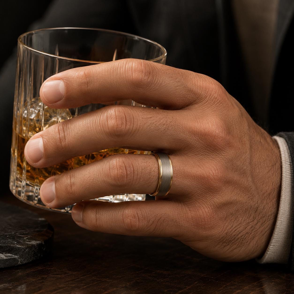 A hand wearing a gold wedding ring is holding a glass of amber-colored liquid, likely whiskey, while sitting at a table.