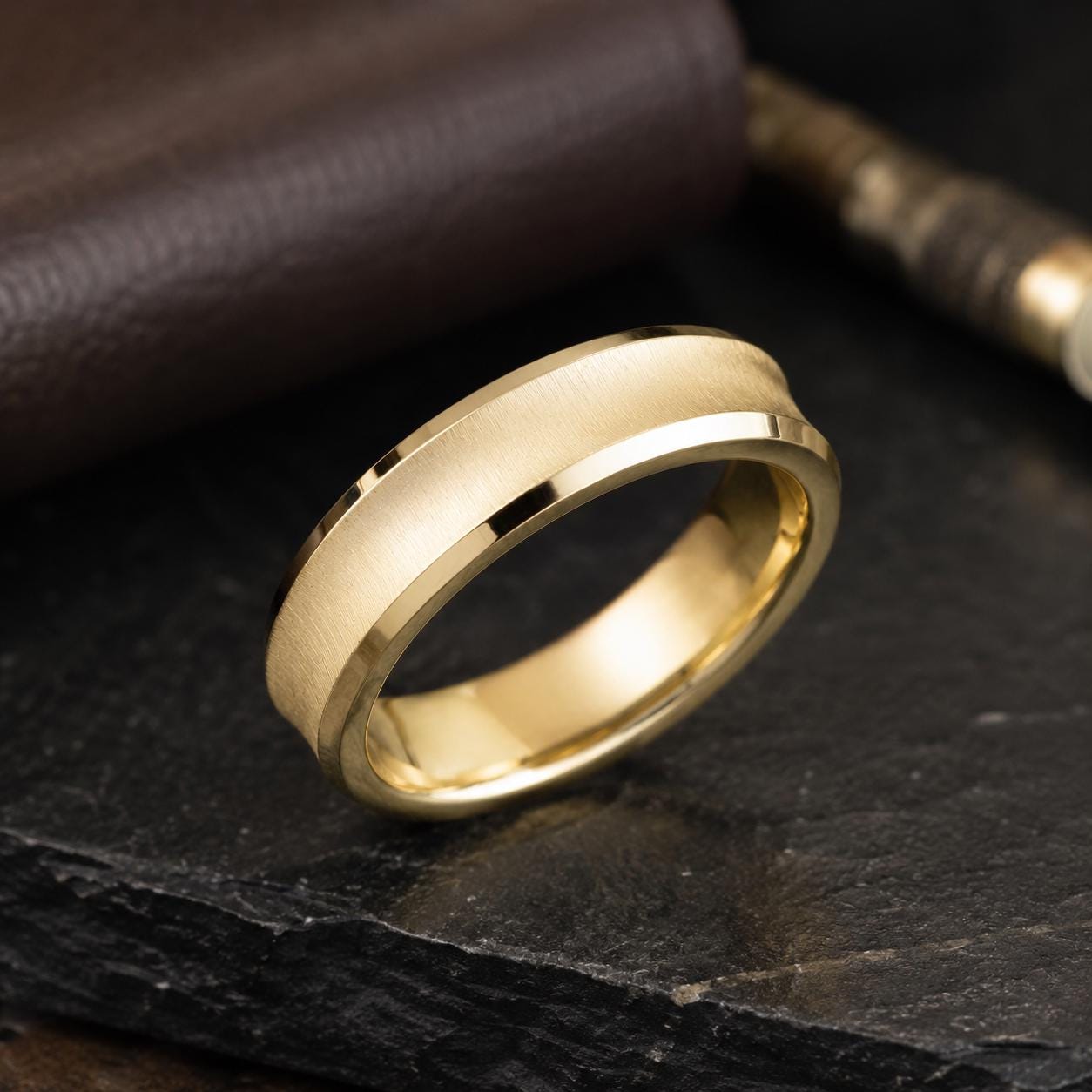 A gold ring with a textured band is displayed on a dark surface, with a brown leather item partially visible in the background.