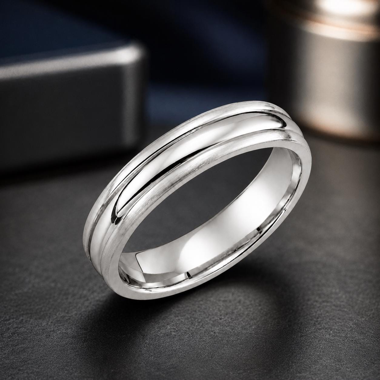 A silver ring with a smooth, polished surface sits on a dark surface, with a blurred background that includes a piece of jewelry and a box.