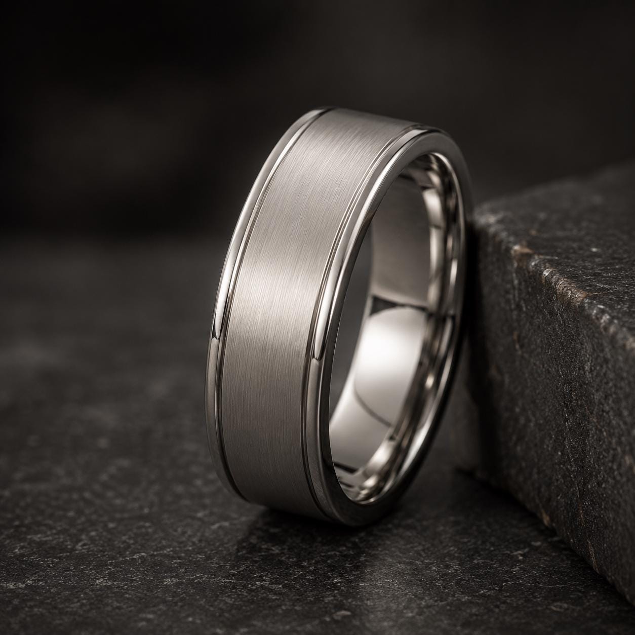 A silver-colored ring with a textured band is resting on a dark surface, such as a stone or a black background.