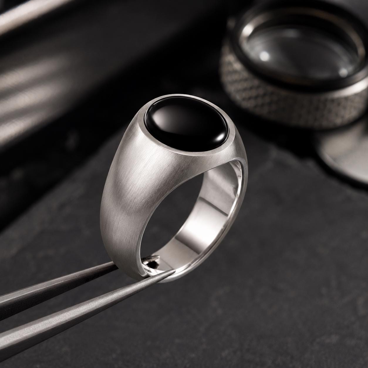 A silver ring with a black gemstone is displayed on a pair of tweezers, with a magnifying glass and a piece of jewelry visible in the background.