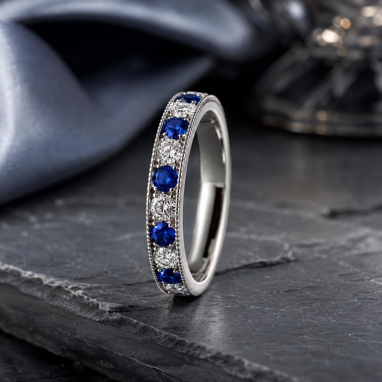 A silver band ring with blue sapphires and diamonds set in a decorative pattern, resting on a dark surface.