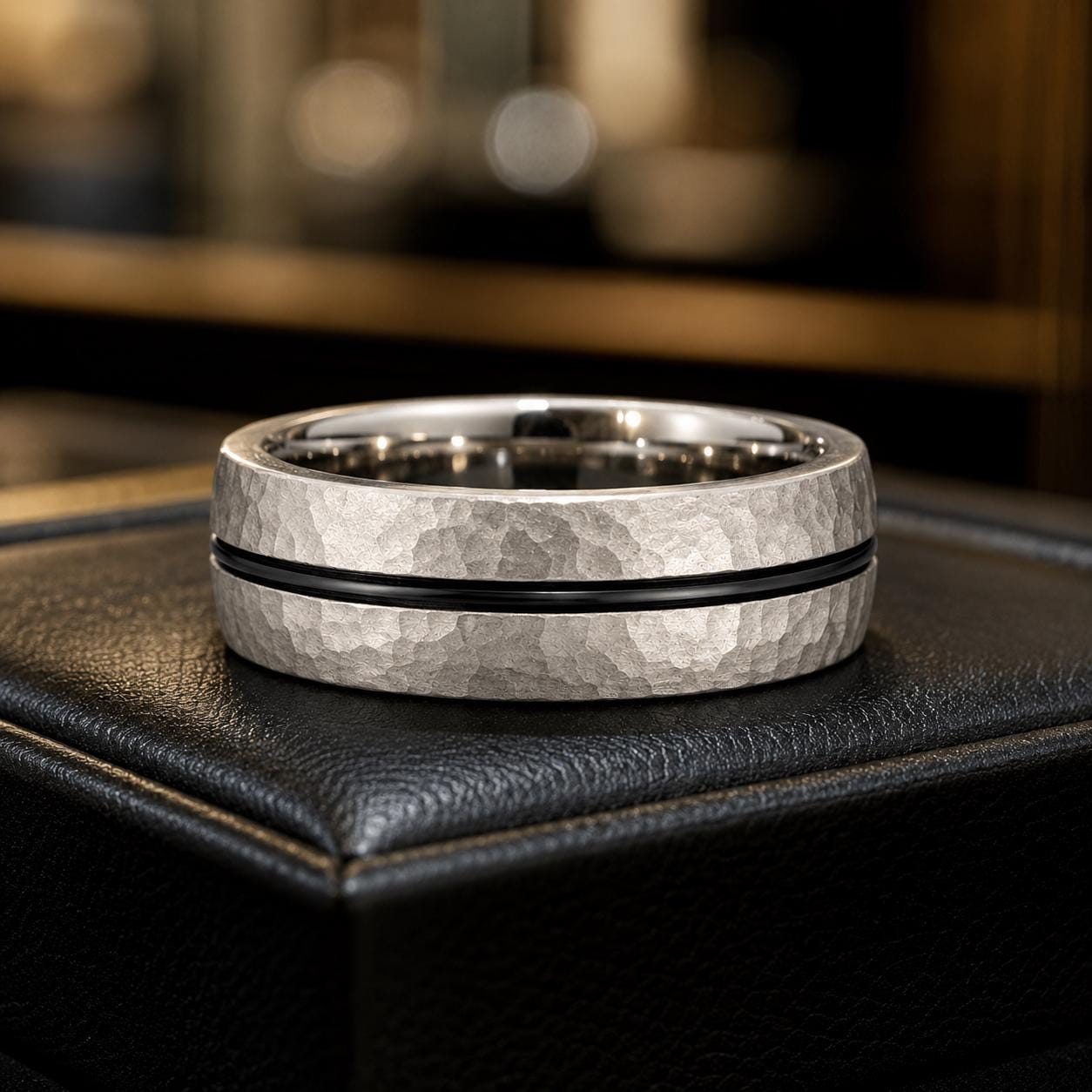 A silver ring with a black band is displayed on a black leather surface.
