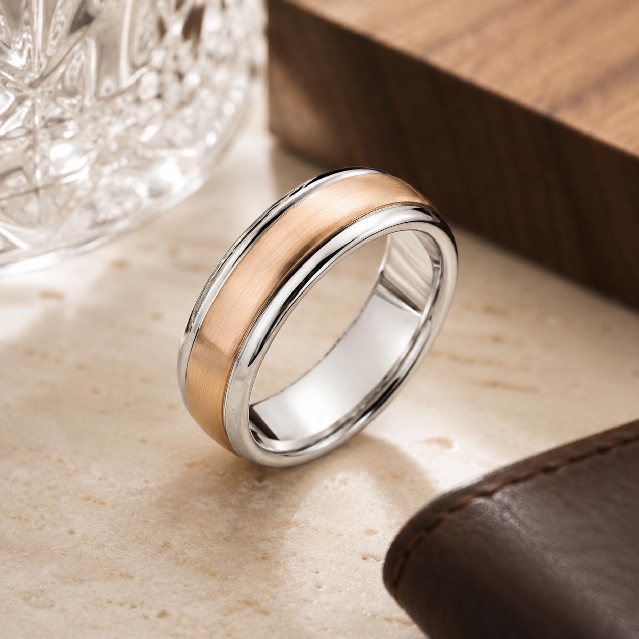 A silver ring with a gold band is displayed on a wooden surface, accompanied by a brown leather wallet and a glass object.
