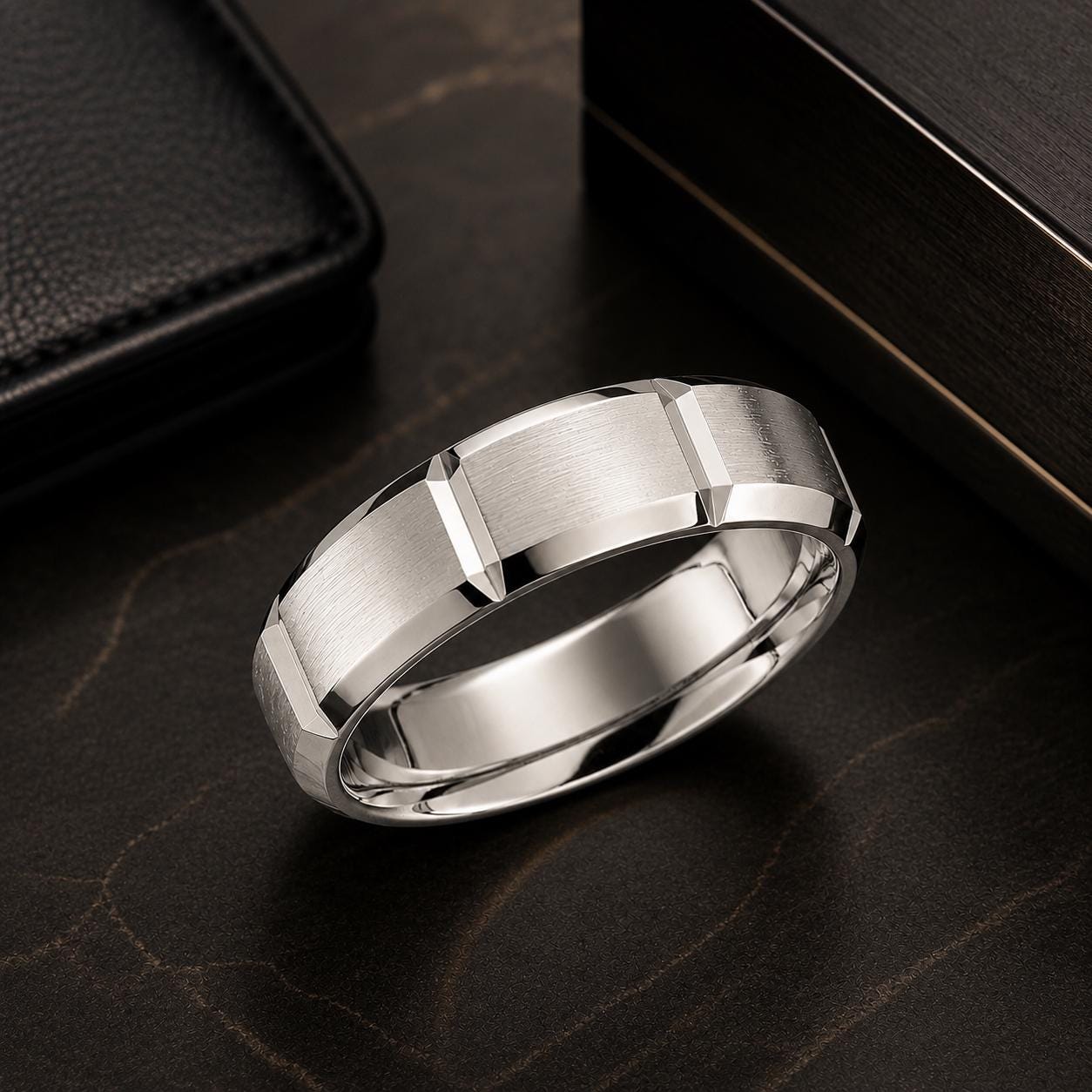 A silver ring with a geometric design sits on a dark surface, accompanied by a black leather wallet and a wooden box.