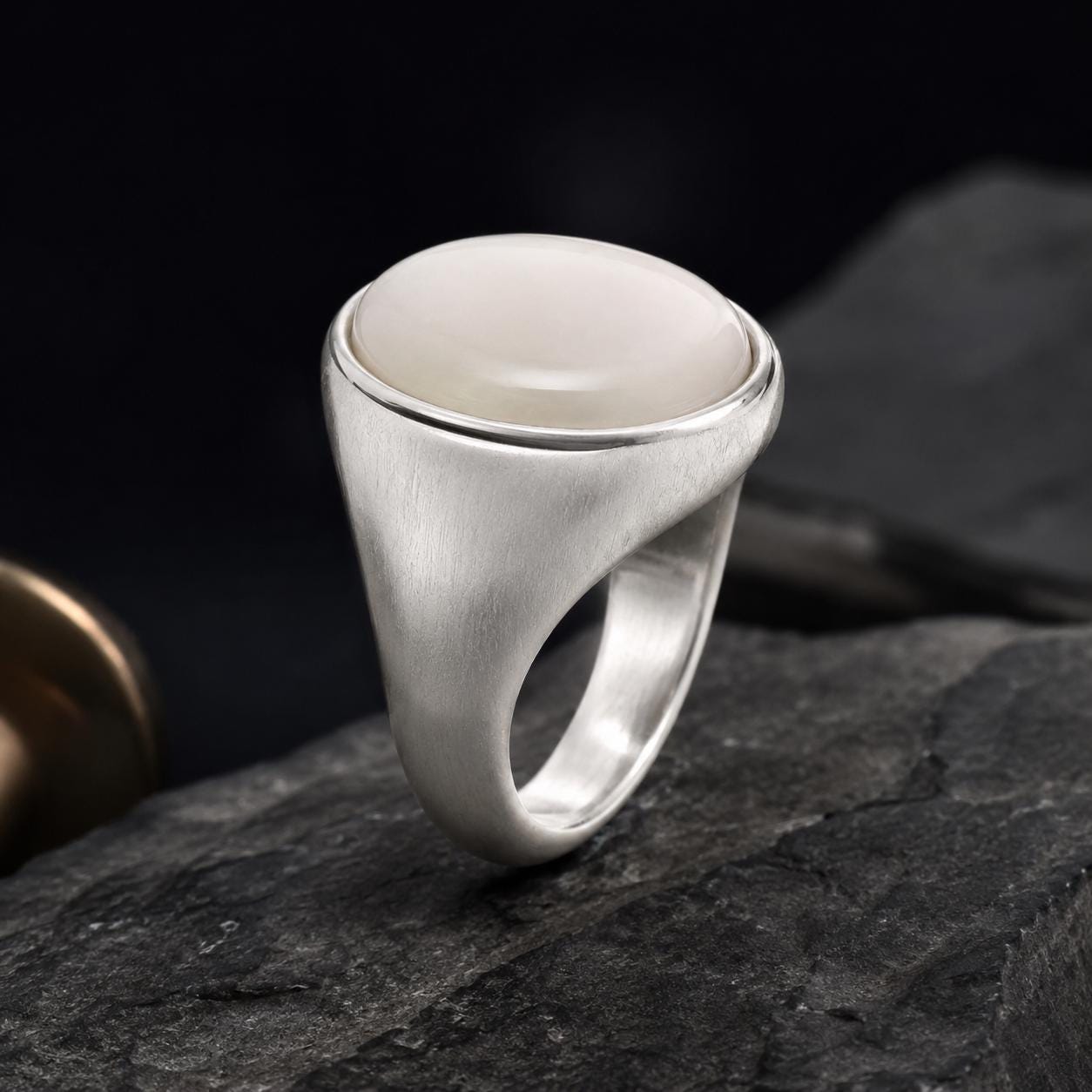 A silver ring with a large white stone sits on a dark surface, possibly a rock.