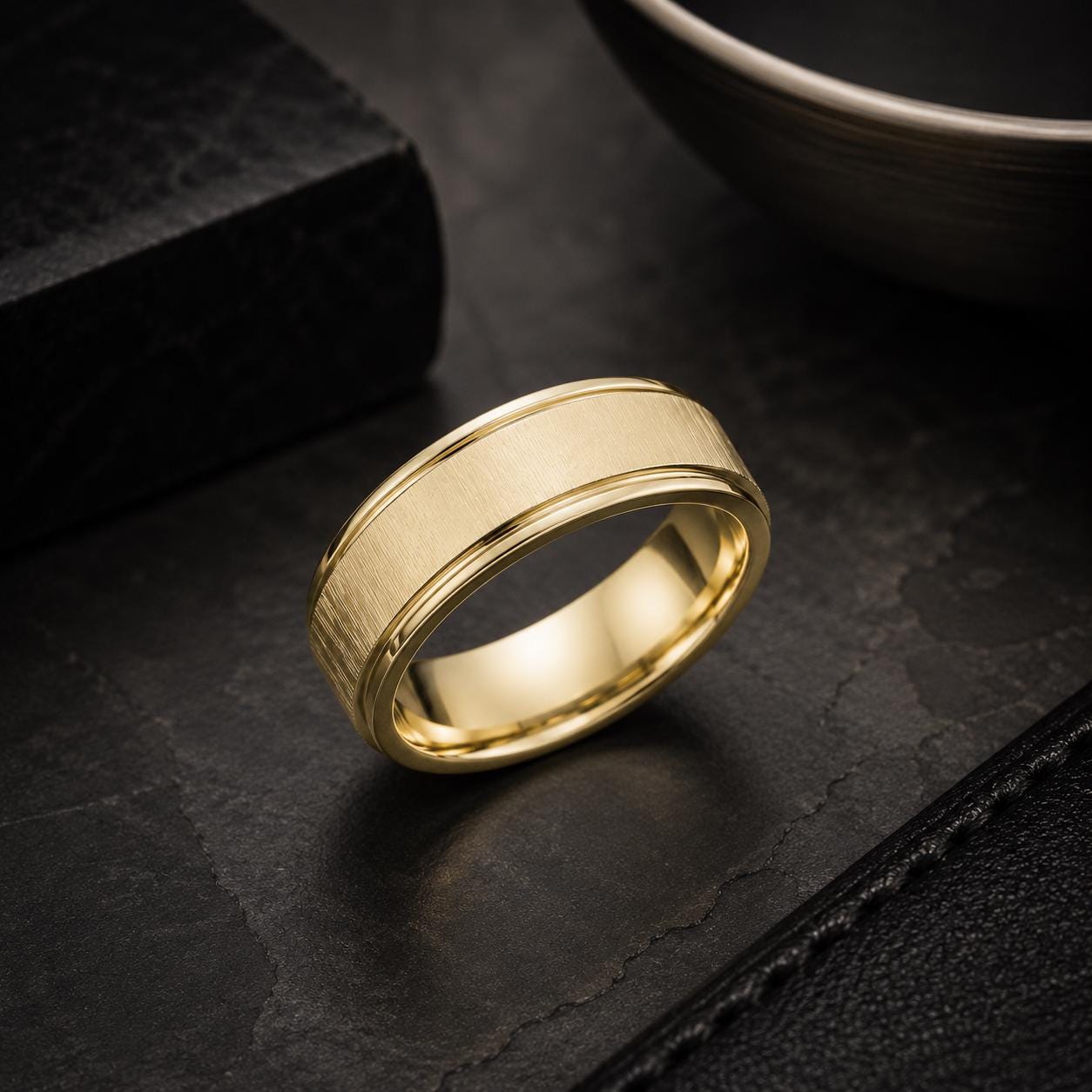 A gold ring with a textured band is displayed on a black surface, with a bowl and a black box in the background.