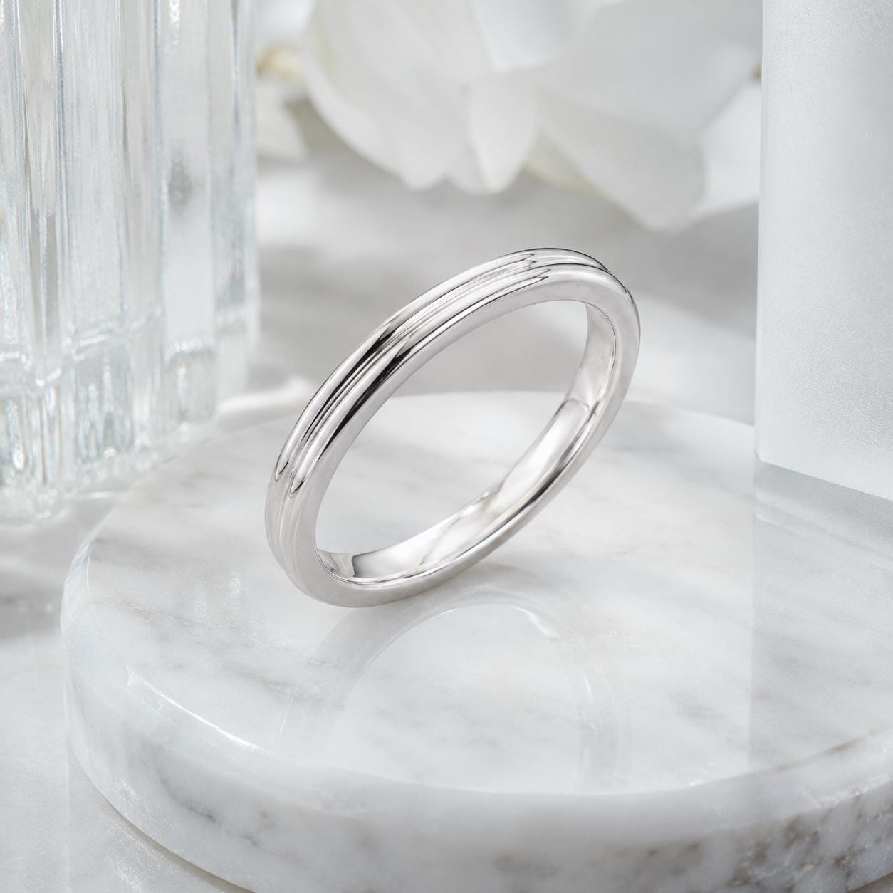 A silver ring is displayed on a white marble surface, with a glass object and white flowers in the background.