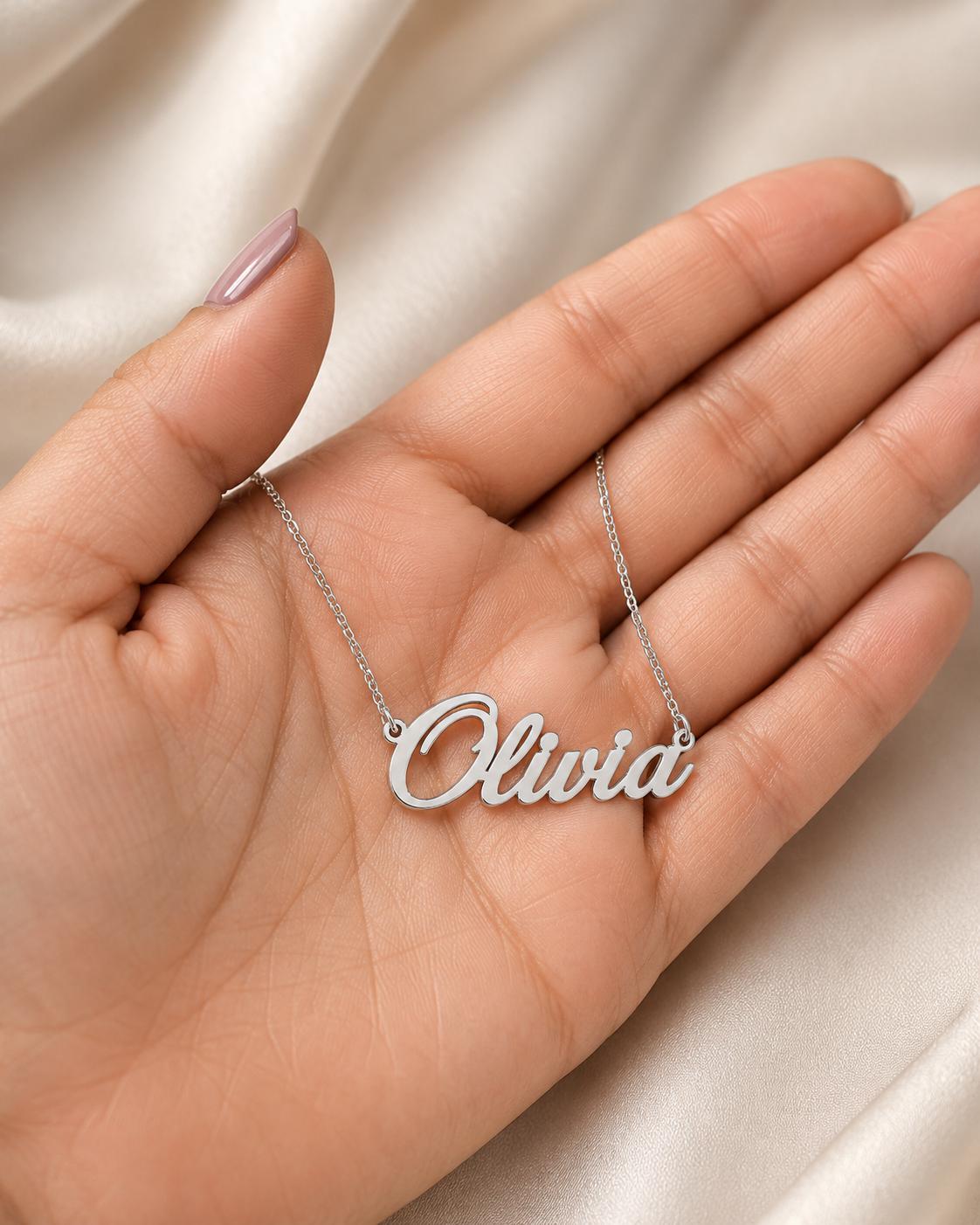 A hand holding a silver necklace with the name "Olivia" engraved on it.