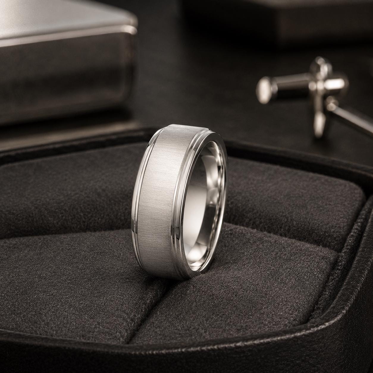 A silver ring with a textured band sits on a black leather surface, with a silver object and a black surface in the background.