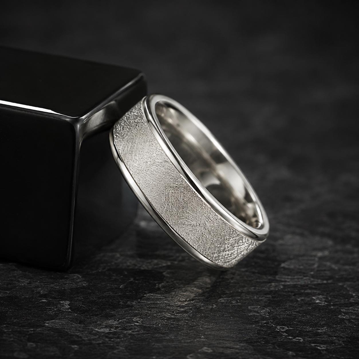 A silver ring with a textured band is placed on a black surface, leaning against a black rectangular object.