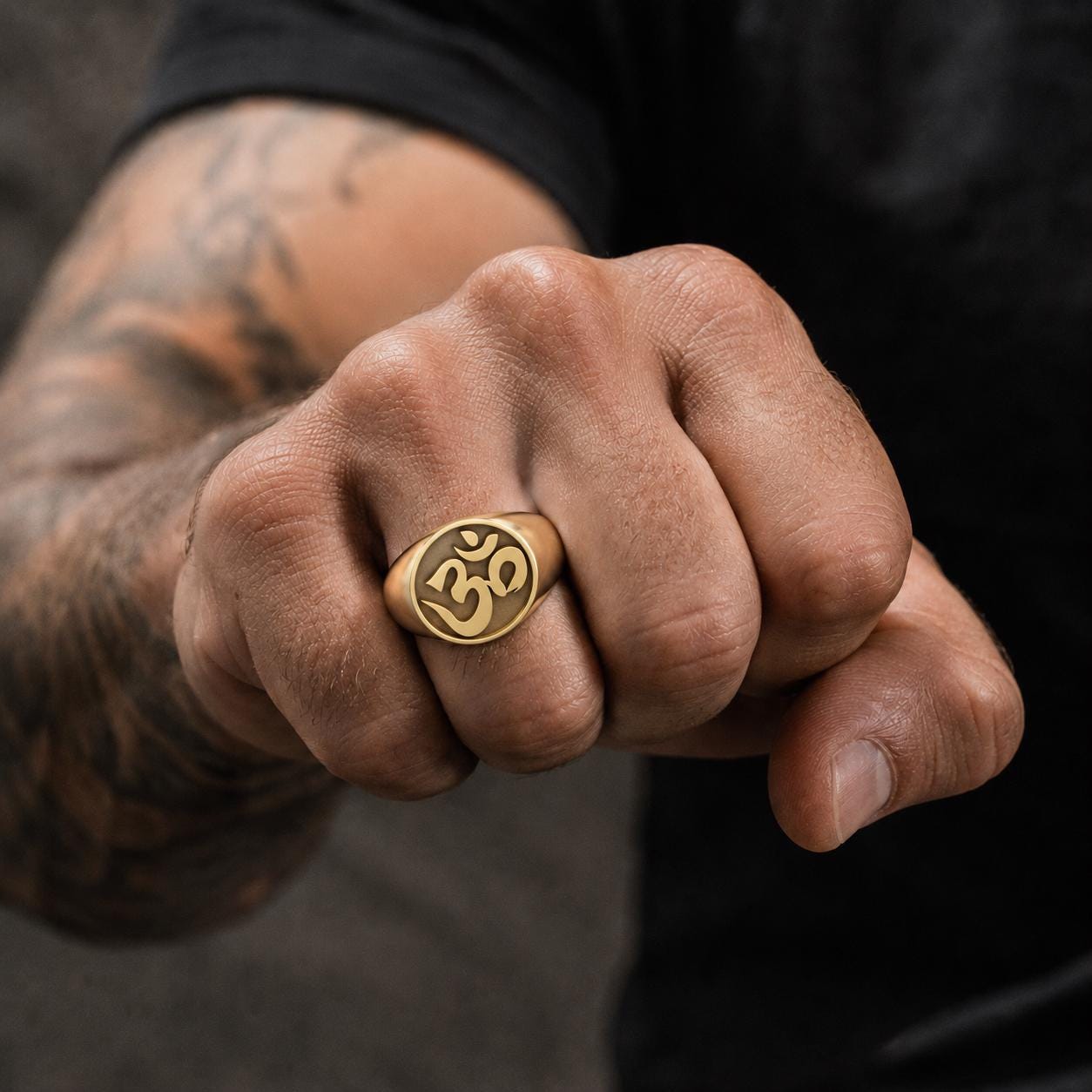 A close-up of a person's hand wearing a gold ring with the Om symbol, with a tattoo visible on the arm.