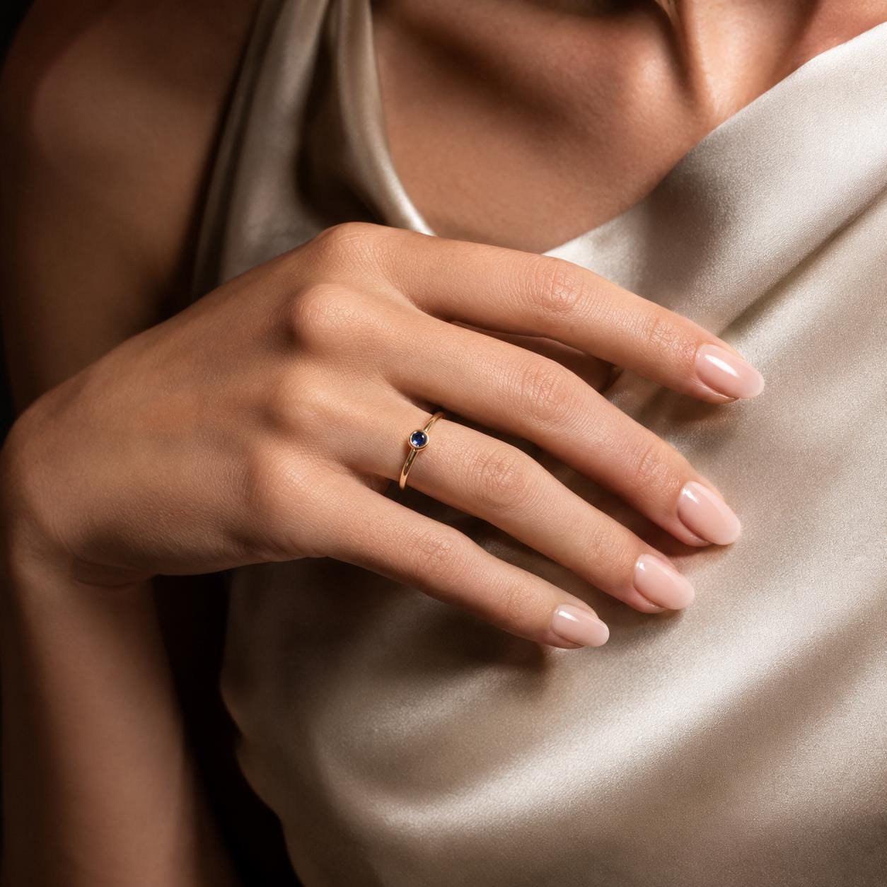 A close-up of a hand wearing a gold ring with a blue gemstone, resting on a beige fabric.
