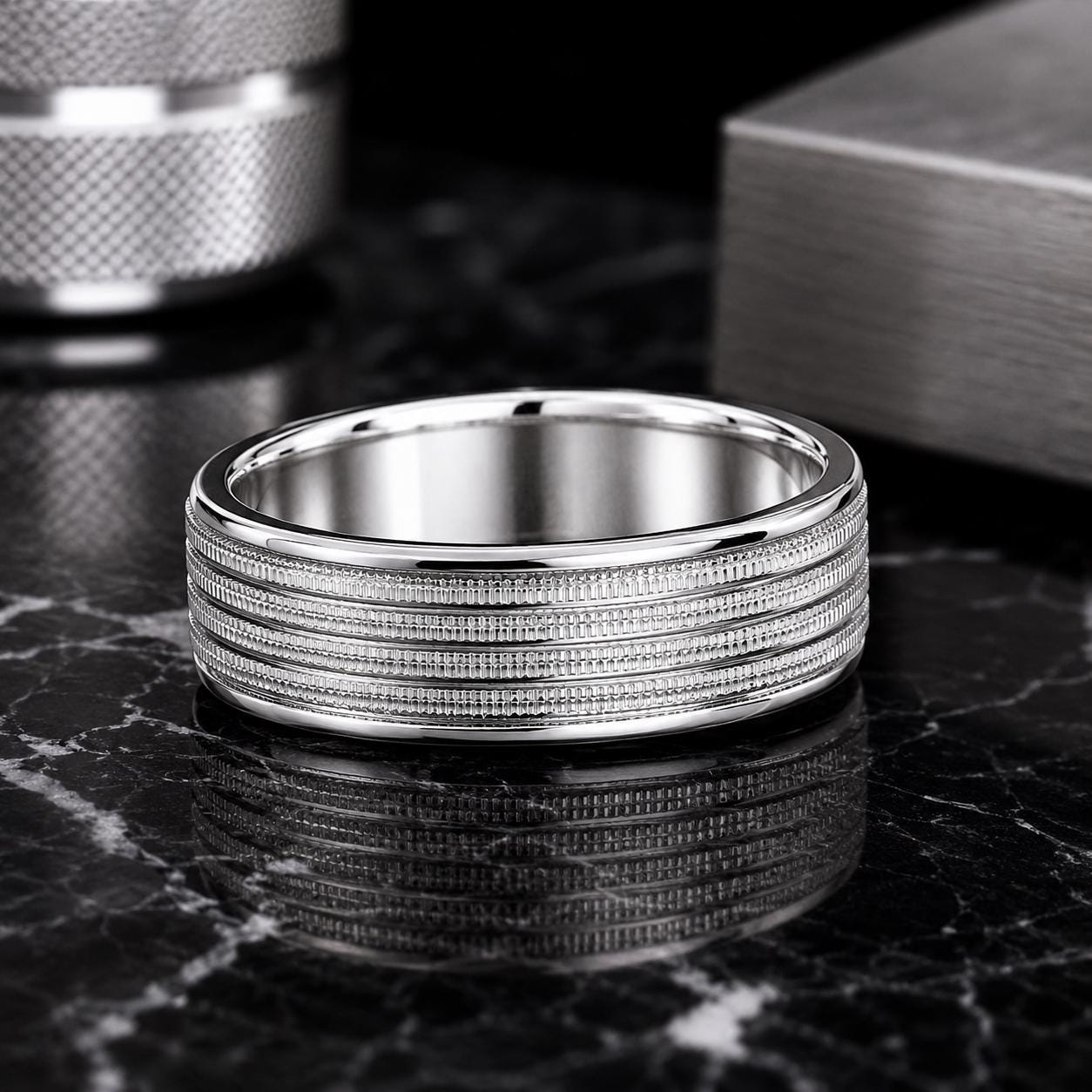 A silver ring with a textured band is displayed on a black marble surface, with a metallic object in the background.