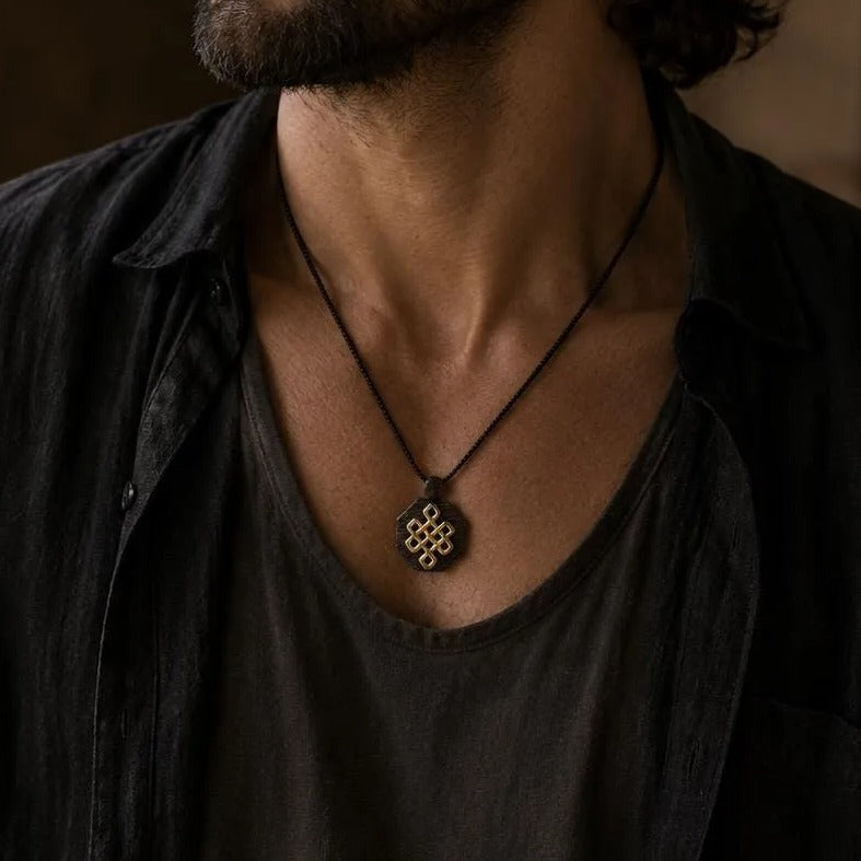 A man with long, dark hair and a beard wearing a black shirt and necklace stands in a room with a window in the background.