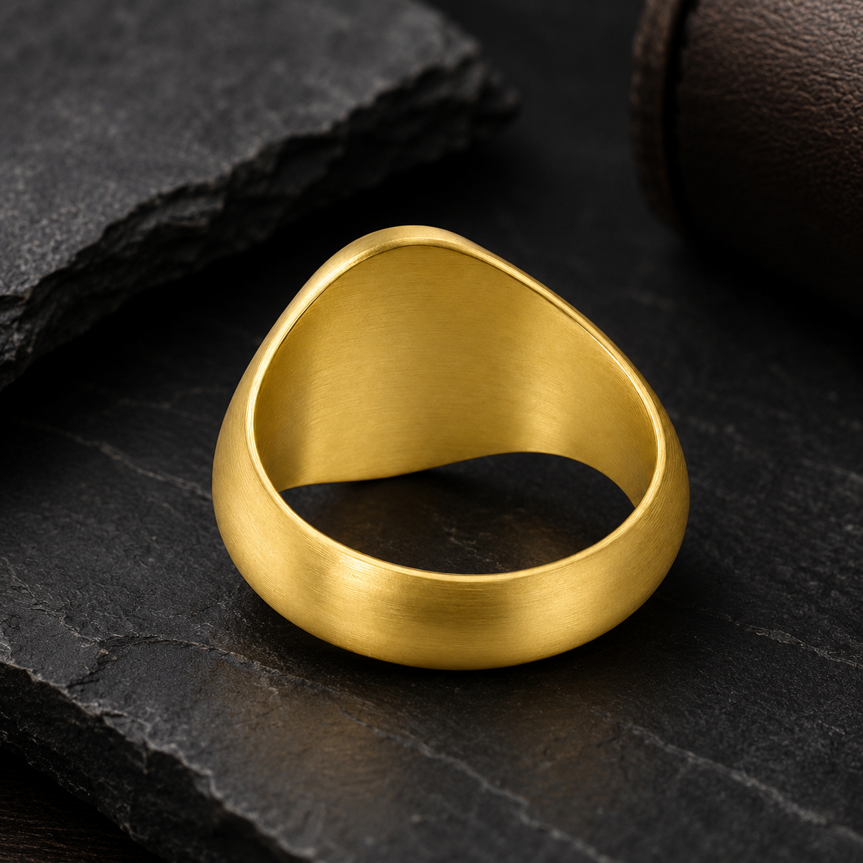 A gold ring with a unique design sits on a dark surface, surrounded by a few other dark objects.