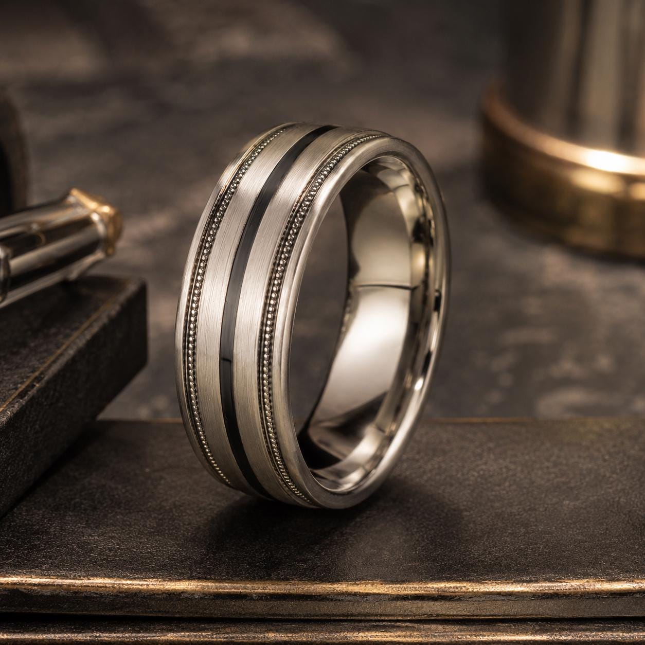 A silver ring with a black band is displayed on a dark surface, with a pen and a piece of paper visible in the background.