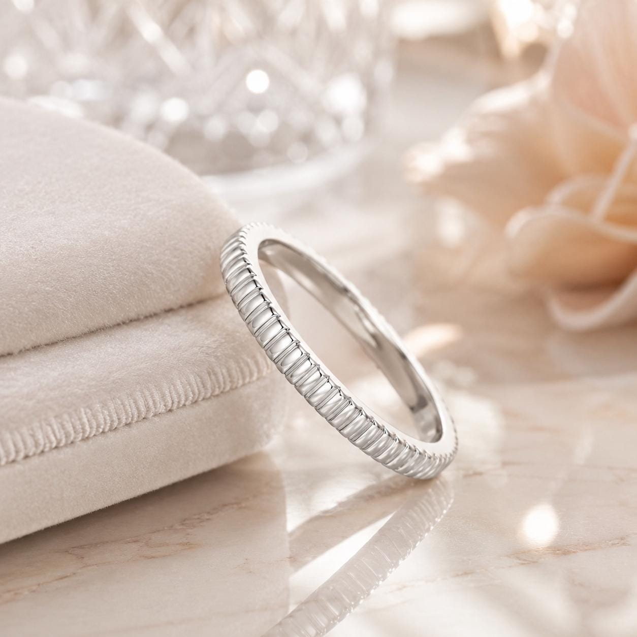 A silver ring with a textured band is displayed on a marble surface, accompanied by a white cloth and a delicate pink flower.