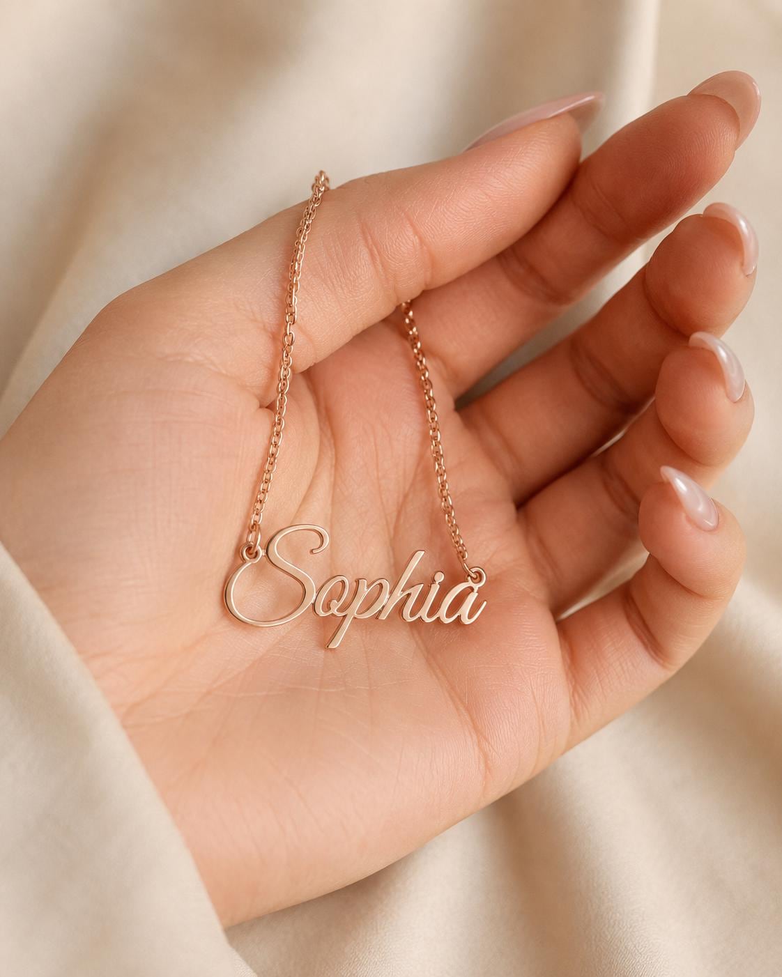 A hand holding a gold-colored necklace with the name "Sophia" engraved on it.