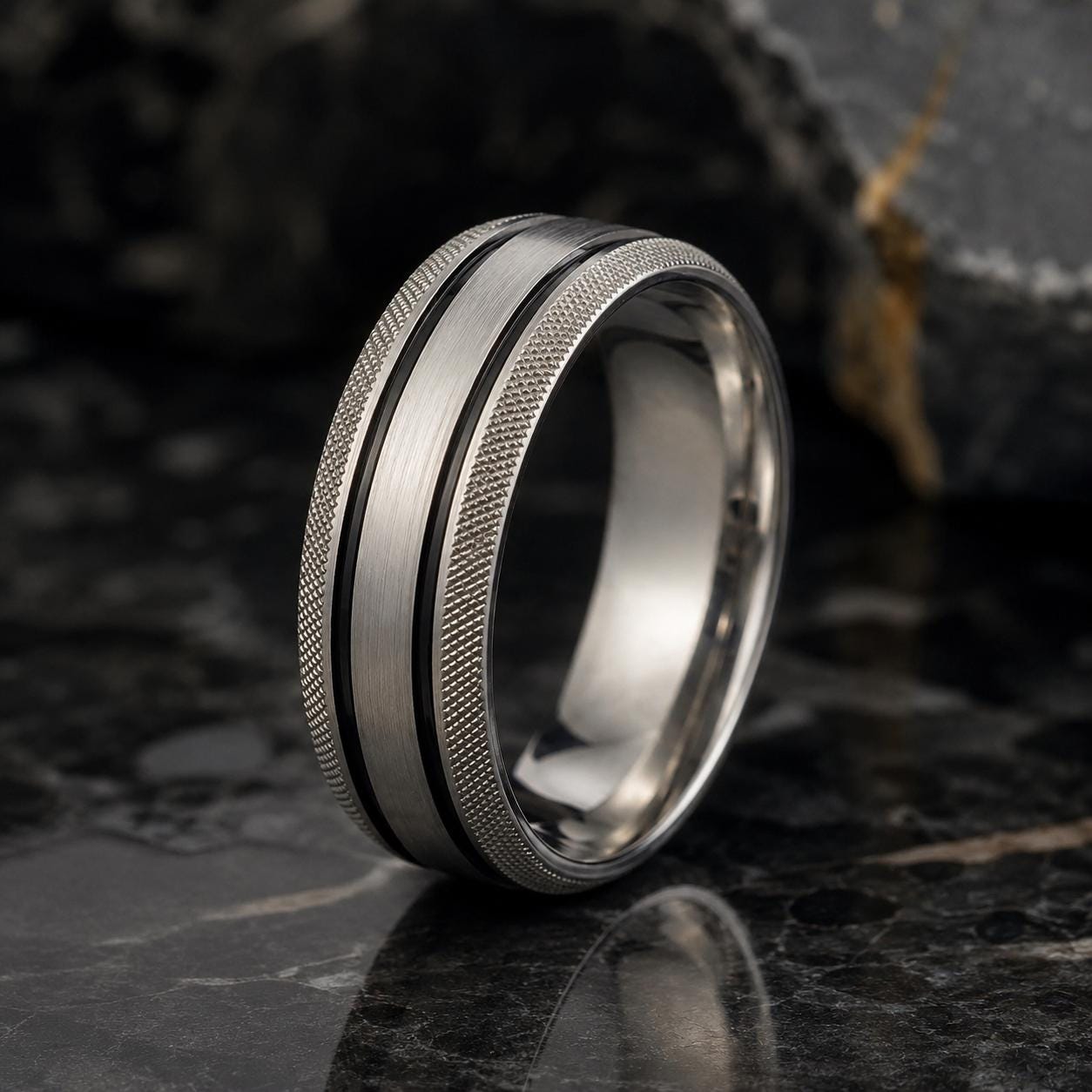 A silver ring with a textured band and a reflective surface, possibly a marble table, is the main focus of the image.
