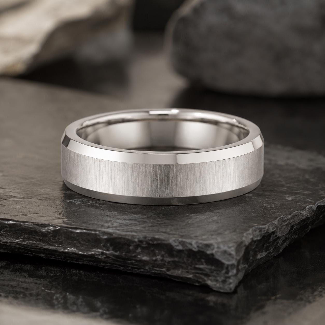 A silver-colored ring with a textured band sits on a dark surface, surrounded by rocks.