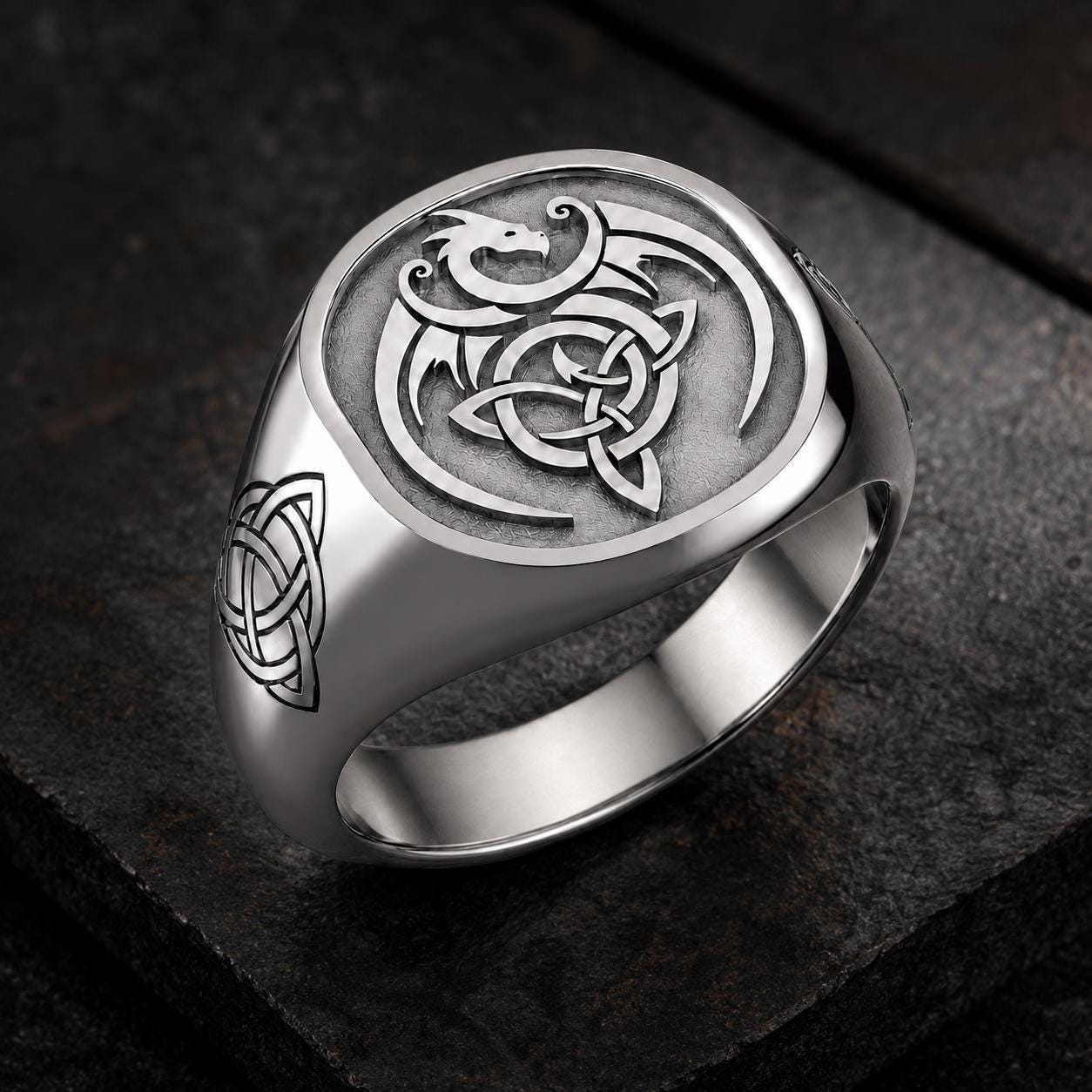 A silver ring with a dragon design on it, sitting on a dark surface.
