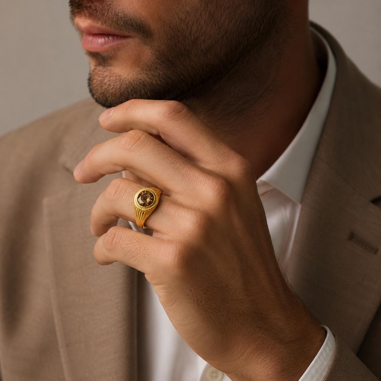 A close-up of a man's hand wearing a gold ring, with a beige jacket and white shirt visible in the background.