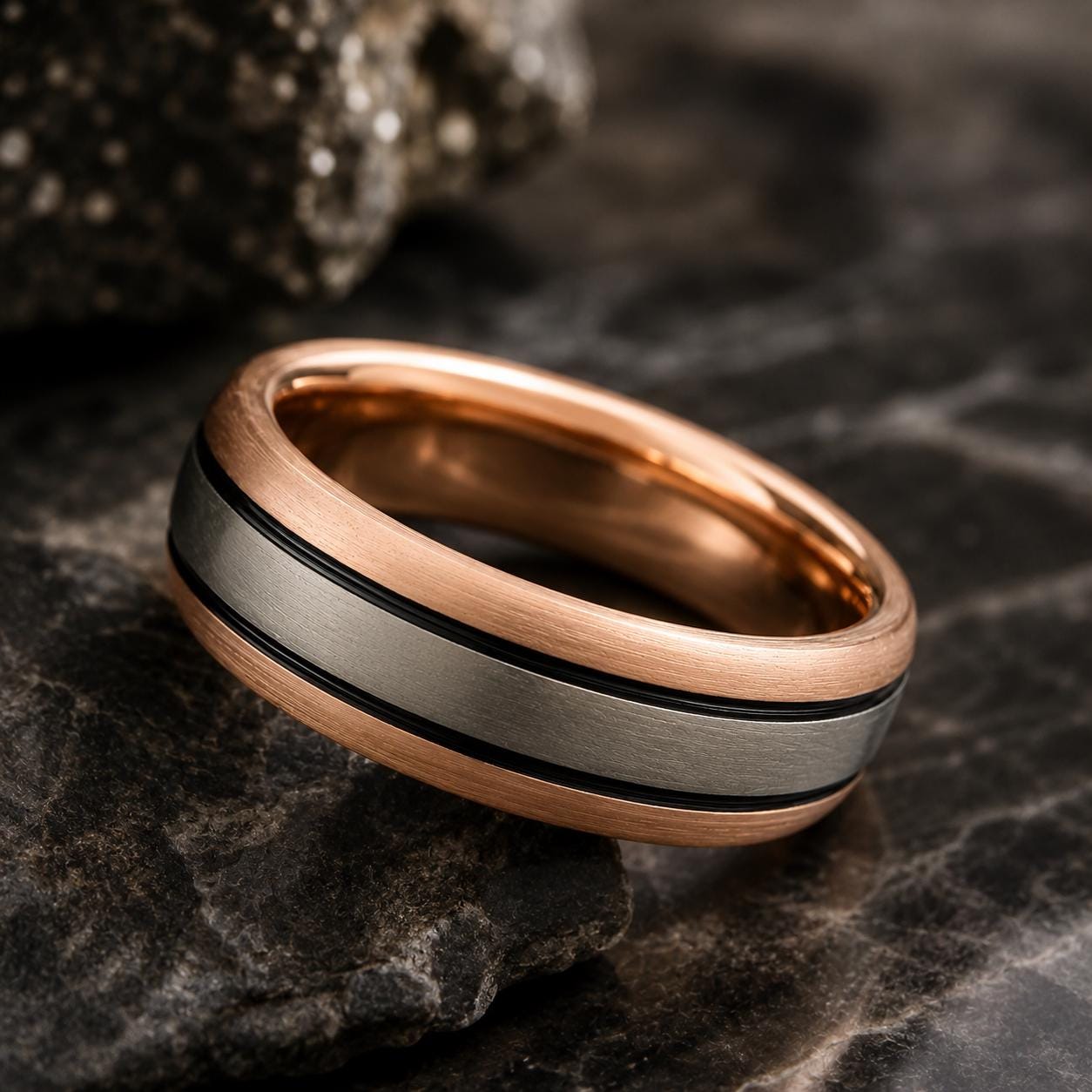 A gold and silver ring with a black stripe is resting on a dark surface, possibly a rock.