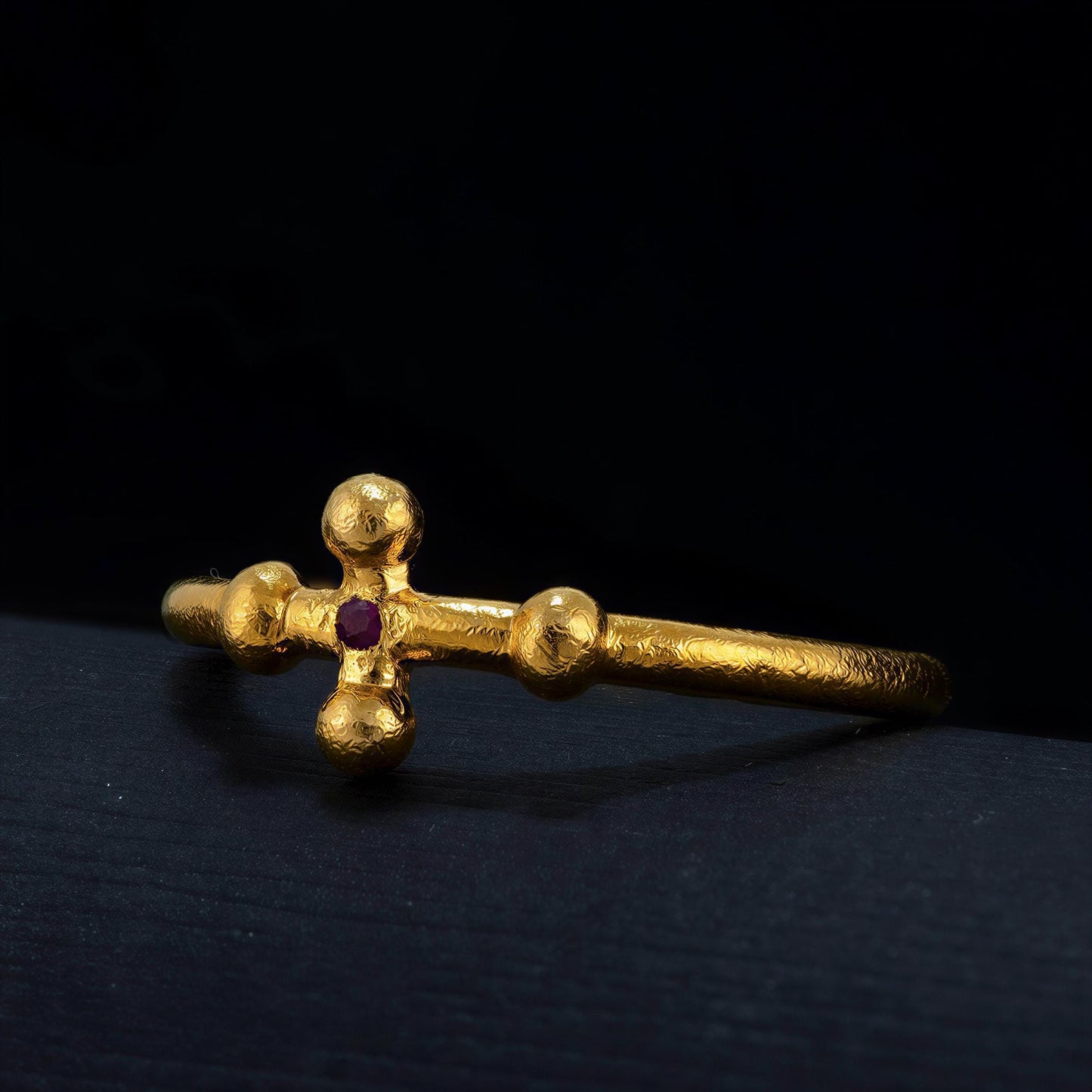 A gold bracelet with a cross-shaped design and a red gemstone is shown against a dark background.