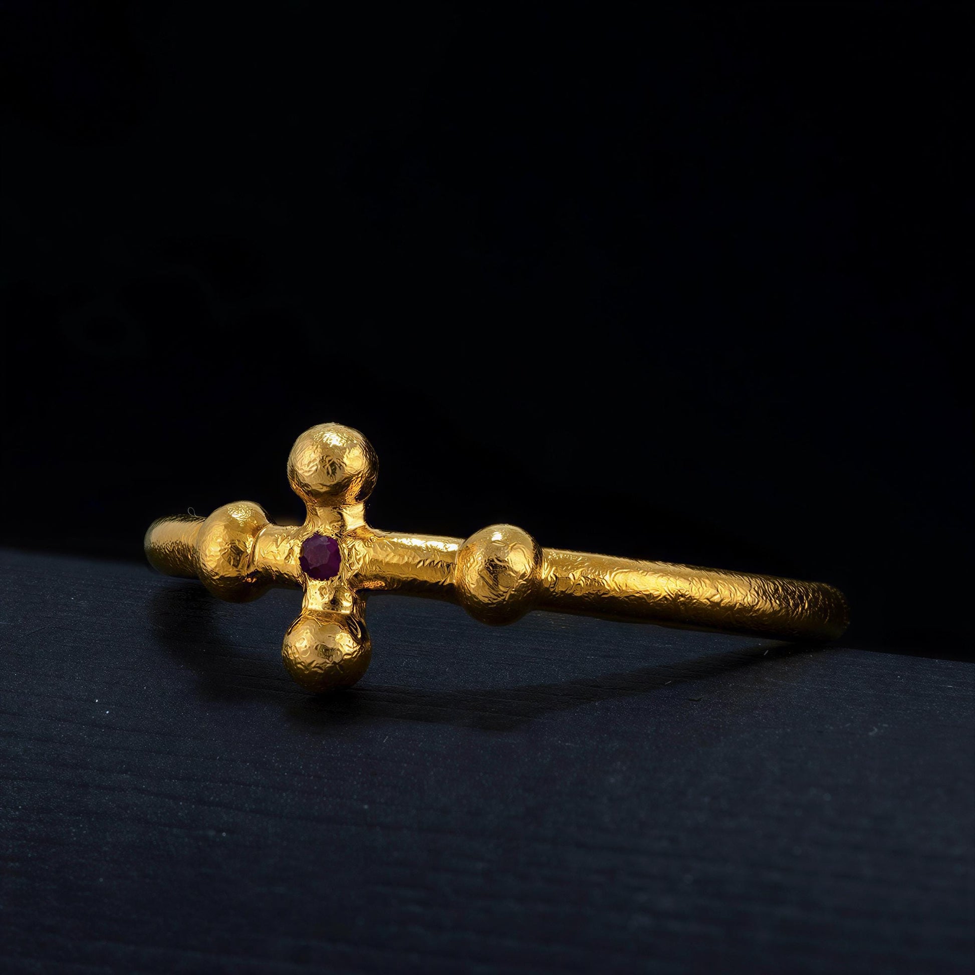 A gold bracelet with a cross-shaped design and a red gemstone is shown against a dark background.