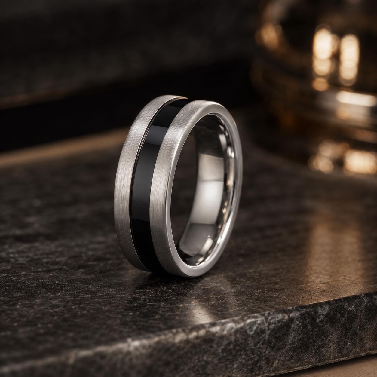 A silver ring with a black band is displayed on a dark surface, with a blurred background.