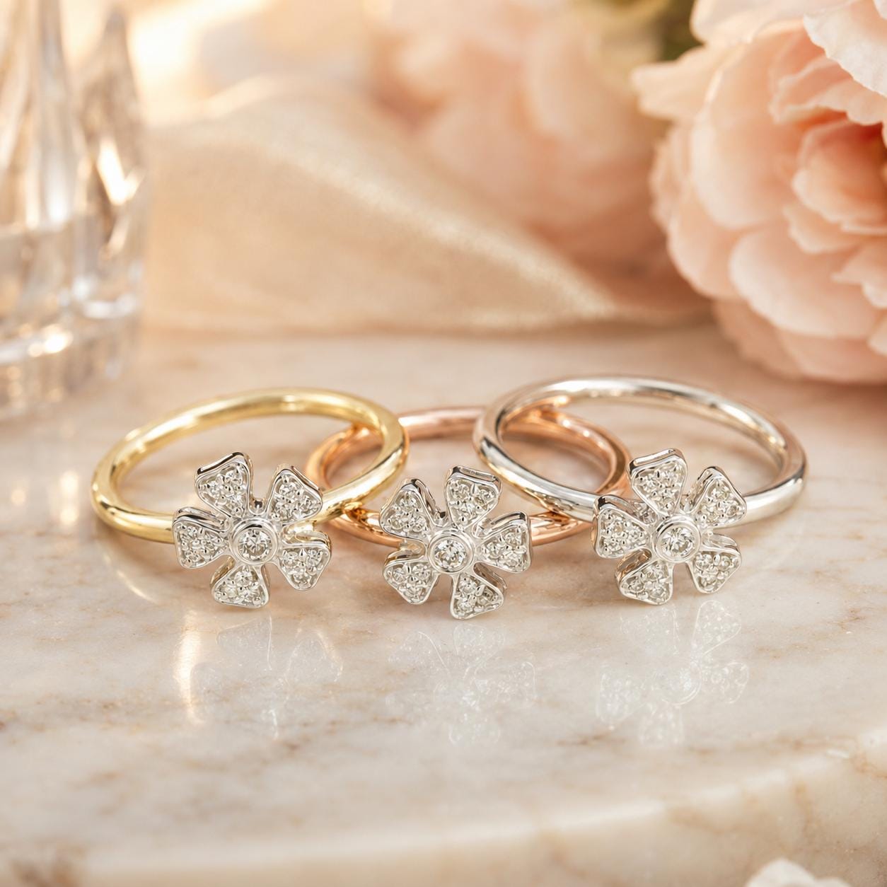 Three diamond-encrusted rings with flower-shaped designs are displayed on a marble surface, with a blurred background of pink flowers.