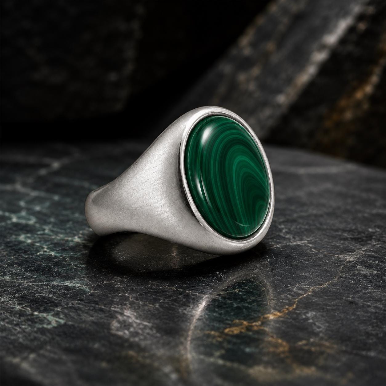 A silver ring with a large oval-shaped green gemstone, likely malachite, is displayed on a dark surface.