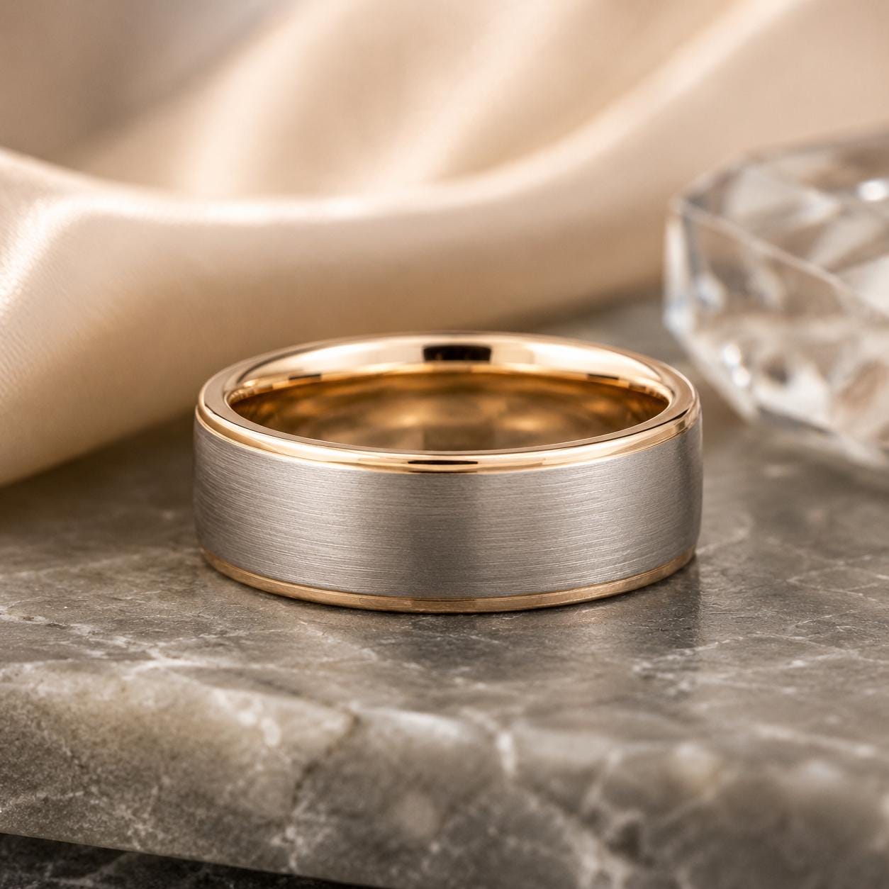 A gold and silver ring with a silver band sits on a marble surface, surrounded by a beige fabric and a clear crystal object.